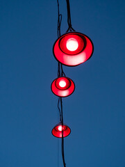 Three lamps hanging on cables in front of a blue evening sky. Red light is shining out of the light...