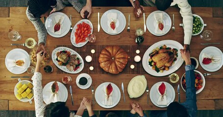 Happy group of diverse religious friends having dinner and holding hands praying over a dining table before eating supper. Overhead view of people having a celebration feast for a special occasion.