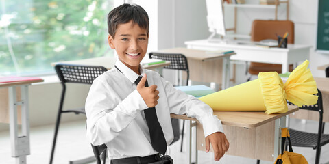 Happy little boy with school cone showing thumb-up gesture in classroom