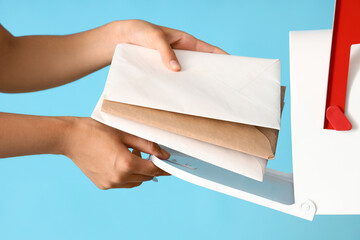 Young postwoman putting letters into mailbox on blue background, closeup