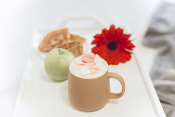 light photo with breakfast, coffee with marshmallows, green apple, red flower and pancakes on a white tray, blurred background, selective focus