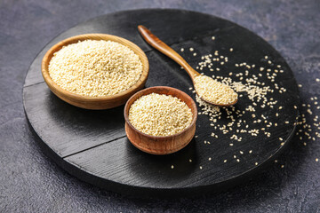 Wooden board with bowls and spoon of sesame seeds on dark background, closeup