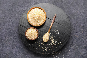 Wooden board with bowls and spoon of sesame seeds on dark background