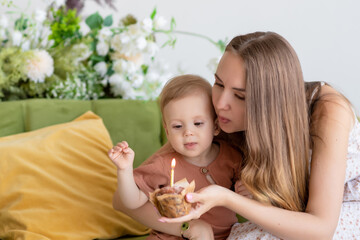 Beautiful loving mother in a dress sits together with her little son on a green sofa surrounded by beautiful flowers. Holds a cupcake with a candle in his hand. Blows out the candle. Festive dessert