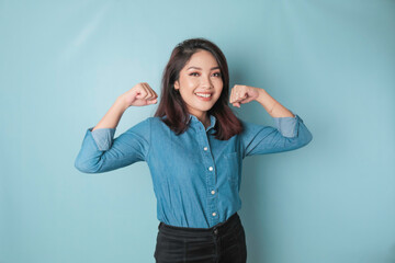 Obraz na płótnie Canvas Excited Asian woman wearing a blue shirt showing strong gesture by lifting her arms and muscles smiling proudly