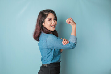 Excited Asian woman wearing a blue shirt showing strong gesture by lifting her arms and muscles...
