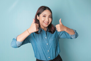 Excited Asian woman wearing a blue shirt gives thumbs up hand gesture of approval, isolated by blue background