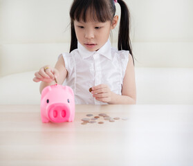 Sweet little preschool girl putting coin into pink  piggy bank at table indoors. Primary economic education