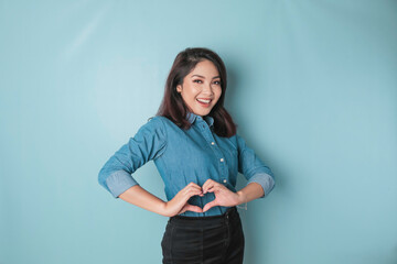 An attractive young Asian woman wearing a blue shirt feels happy and a romantic shapes heart gesture expresses tender feelings