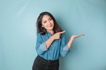 Excited Asian woman wearing blue shirt pointing at the copy space beside her, isolated by blue background