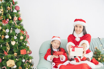 Fototapeta na wymiar Asian mother and daughter in Santa suit with gift box sitting in room decorated with Christmas tree
