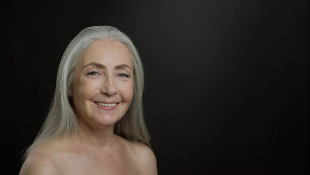 Beautiful senior lady turning face to camera and smiling, posing with bare shoulders, black background, empty space