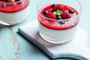 Panna cotta dessert with sauce from berries. Cherries, raspberries and blueberry as decoration. Served in two glasses on dark background. Traditional Italian creamy dessert.