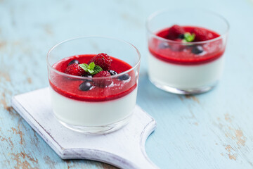 Panna cotta dessert with sauce from berries. Cherries, raspberries and blueberry as decoration. Served in two glasses on dark background. Traditional Italian creamy dessert.