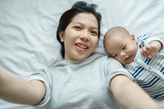 Asian Young mother and little baby son lying on bed having video call with family with smartphone or mobile phone together