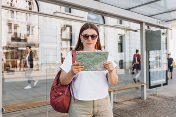 A cheerful caucasian redhead woman of an urban bus stop and using paper map. Urban travel and transportation concept. Girl search location in the city