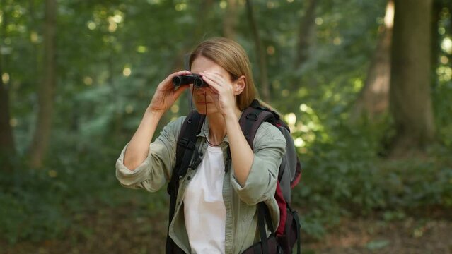 Middle aged active woman tourist peering into distance with binoculars, hiking with backpack in forest, empty space