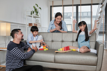 Fototapeta premium Happy Asian lovely Thai family care, dad, mum, and little children have fun playing with colorful toy blocks together on sofa in white living room, leisure weekend, and domestic wellbeing lifestyle.