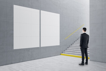 Young businessman standing in creative light concrete tile interior with stairs and mock up poster on wall. School hallway and corridor concept.