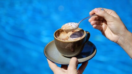 Against the background of blue water, female hands hold a cup of delicious coffee with foam, slow...