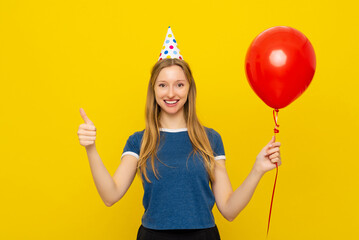 Young positive woman in a birthday cap holds red inflated helium balloon and shows thumb-up, feels very happy, stands over yellow background. Holiday and party concept