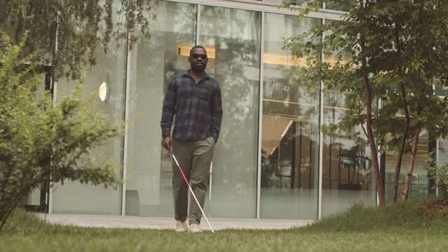 Full length slowmo of young African American blind man in black eyeglasses walking along green grass in city park using walking stick