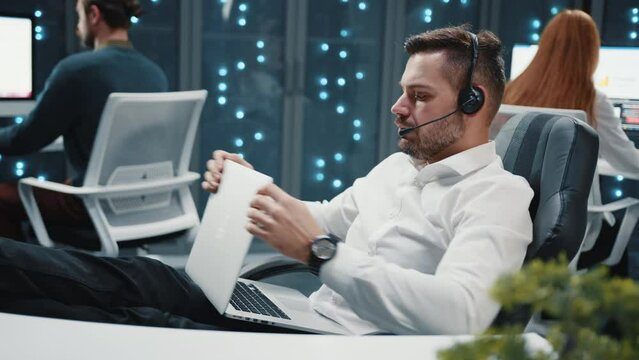 Close up confident attractive young bearded man talking via video call with feet on desk in modern cozy office. Ending conversation thinking. Slow motion