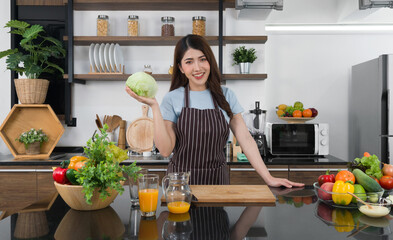 Young asian housewife dressed in an apron, hold fresh lettuce with one hand. The kitchen counter full of various kinds of vegetables.