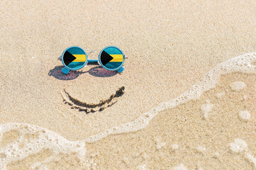 Fototapeta na wymiar A painted smile on the beach and sunglasses with the flag of the Bahamas. The concept of a positive holiday in the resort of the Bahamas.