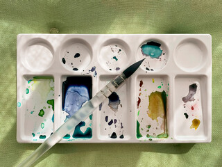 The brush is on the white palette for mixing colors. The paints are splattered in spots and colored blobs. Concept of painting with paints, creativity, hobby.