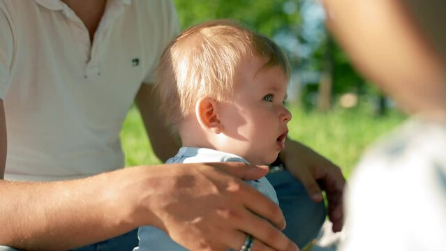 Baby boy sits near father pouting cheeks. Father strokes son with large hand. Toddler child examines park territory with careful and amused expression closeup