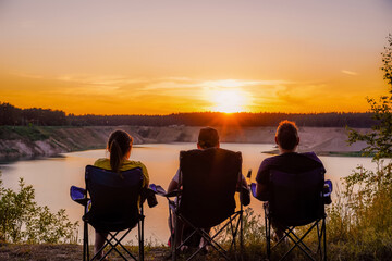 Three young people in armchairs watch the sunset over the lake. Silhouettes of people.