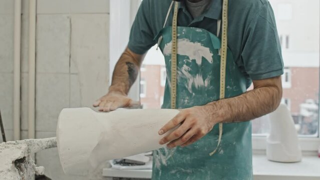 Young adult bearded craftsman wearing apron polishing plaster cast for leg prosthesis socket with sandpaper