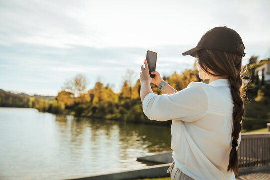 Woman with cap photographing lake through smart phone on sunny day