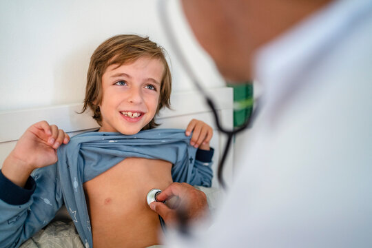 Doctor with stethoscope examining smiling boy in bed at home
