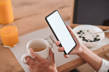 Hands of woman holding coffee cup and smart phone at home