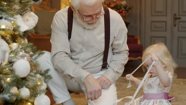Cute little girl opening gift box with assitance of granddad and taking garland from it on Christmas Eve at home