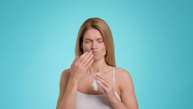 Sick middle aged woman feeling unwell, suffering from runny nose, blowing into paper issue, blue background, slow motion