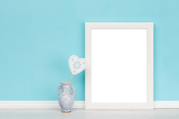 Poster mockup with vase, heart shaped ornament and white wooden vertical picture frame with transparent background in front of turquoise wall.