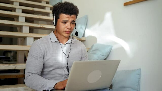 Man in headset speaks at online teaching conference via laptop zoom room sitting on wooden stairs at home. Young guy turns looking in camera
