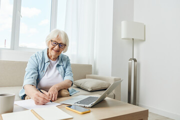 an elderly woman in stylish clothes is sitting on a sofa in a comfortable bright apartment and working on a laptop makes notes in a notebook. The concept of working from home