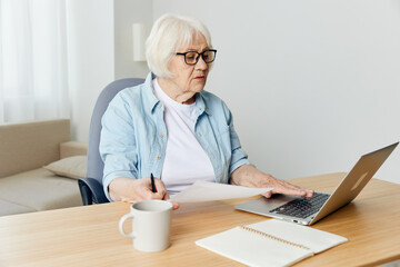 an elderly woman sitting at a laptop and working, from home in her cozy apartment, with a cup and notebooks on the table