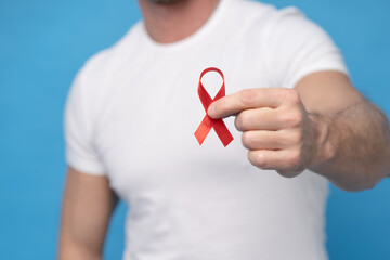 Man with red ribbon bow AIDS awareness symbol in hand wearing a white t-shirt isolated on a blue background. Modern medicine and healthcare. AIDS awareness concept. No face visible