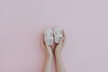 White cotton cute baby shoes in women's hands on pastel pink background. Fashion Scandinavian baby...