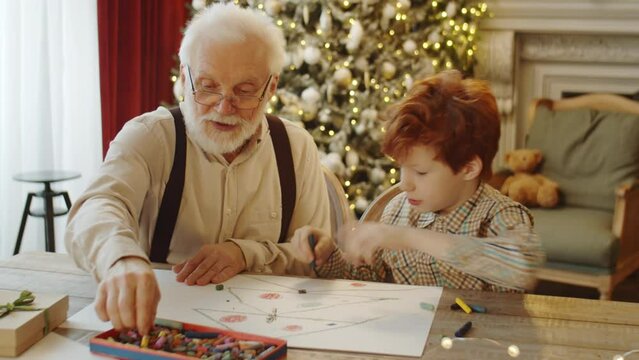 Little boy and elderly grandfather drawing Christmas tree with wax crayons on paper while preparing for holiday at home