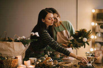Two millennial women making Christmas wreath using pine branches and festive decorations. Small business © polinaloves