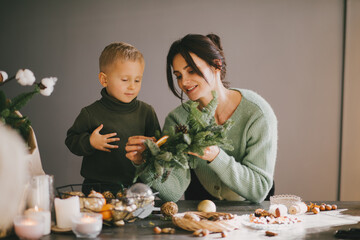 Mother and her little son making Christmas wreath together using pine branches and cones. Festive holiday cozy home decorations.