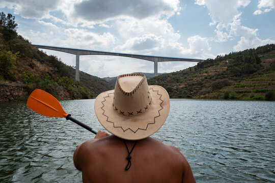 Jovem desportista de chap&eacute;u a praticar desporto com o seu caiaque no rio com uma ponte ao fundo num dia nublado
