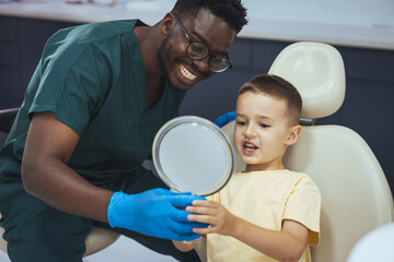 Smiling little boy holding a mirror and looking at his teeth after dental examination at dentist's office. Cute boy looking his teeth in a mirror after dental procedure at dentist's office.