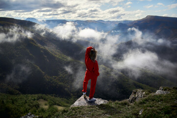 Fototapeta premium A young girl in red stands on the edge of a cliff in the mountains and looks at the clouds that have descended to the Alpine meadows